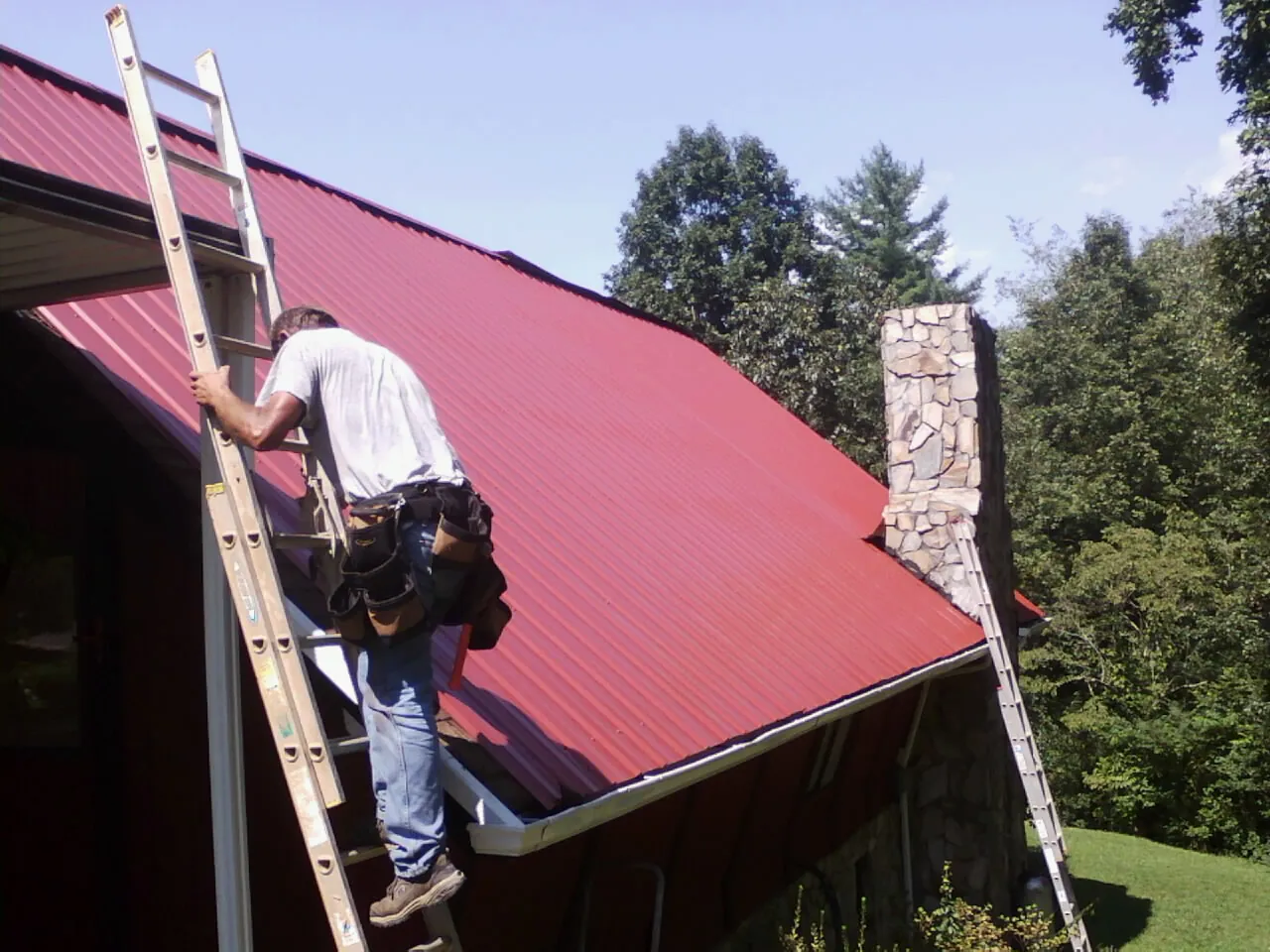Roofer on a ladder beside a red metal roof and stone chimney — Soffit & Fascia Repair by Roofing Brothers in El Reno